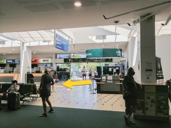 People standing inside the Sunshine Coast Airport looking towards the information desk with an yellow arrow pointing to the left.