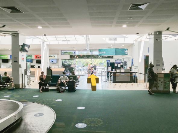 People sitting inside the Sunshine Coast Airport. Facing the exit with a yellow arrow pointing straight in the direction to leave.