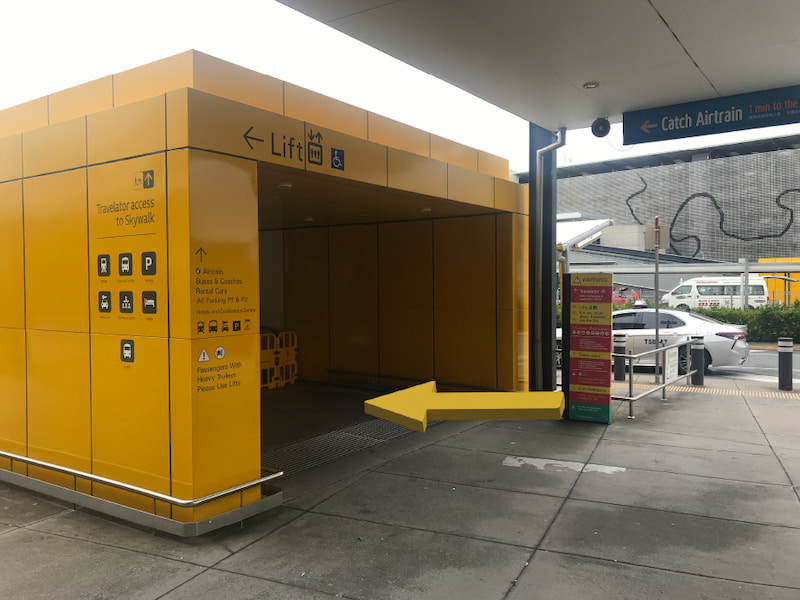 Yellow arrow indicating to use the lift to access the Skywalk. Taxi and airport carpark in the background