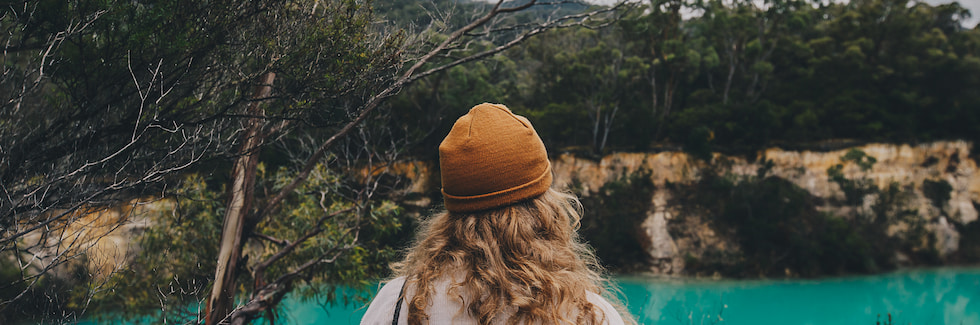 Person in orange toque surrounded by the forest over looking a blue lake.