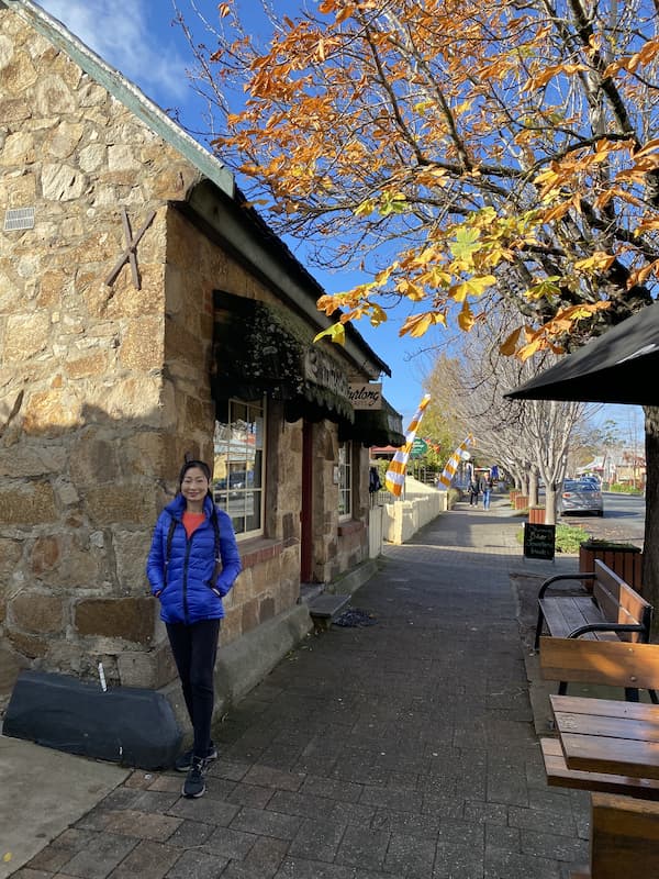 Lady in a blue jack standing beside brick building. Clear blue skys and orange leaves fall off a tree on the right.